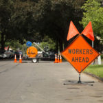 Orange and Black "Workers Ahead" Sign in Residential Neighborhood with Equipment in Background, Daytime