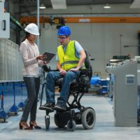 Female manager training a new employee with a disability for a job, explaining work on machines. Industry, inclusion and workplace concept.